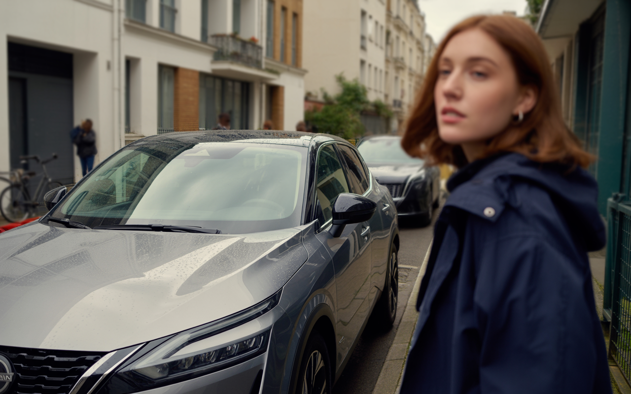 Une jeune femme au cheveux bouclés passe devant une voiture grise sous la pluie, dans une rue résidentielle.