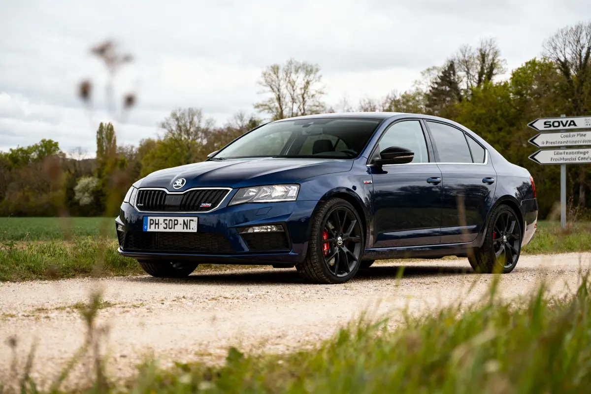 Voiture bleue Škoda sur chemin de campagne, arbres et panneau de signalisation en arrière-plan.