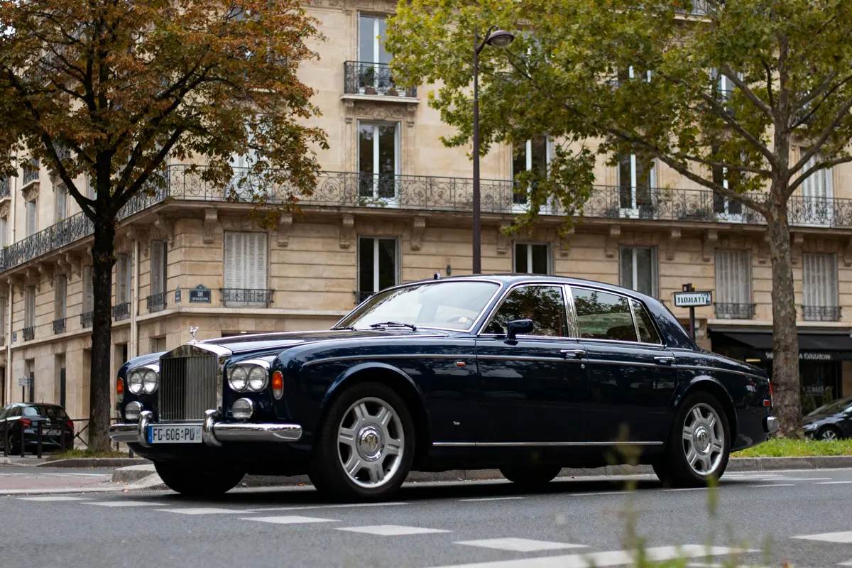 Voiture de luxe noire stationnée devant immeuble parisien, arbres en arrière-plan, ambiance urbaine élégante.