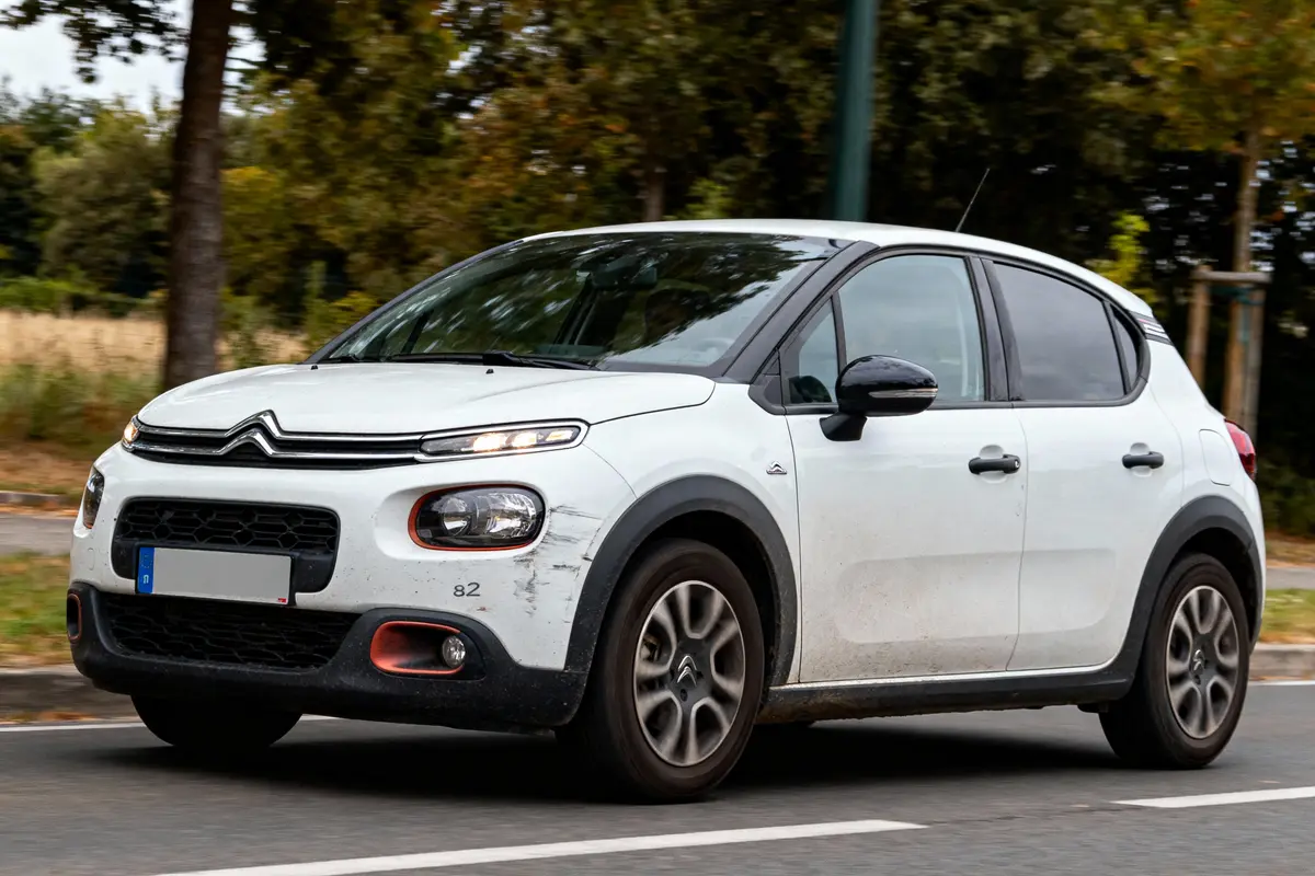 Petite voiture blanche Citroën C3, roulant sur route bordée d'arbres verts, en mouvement vers la gauche.