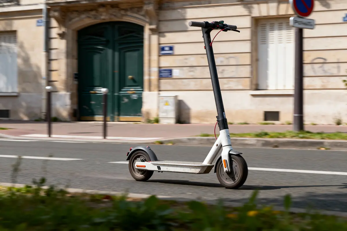 Trottinette électrique blanche sur route, devant bâtiment ancien avec porte verte et panneaux.