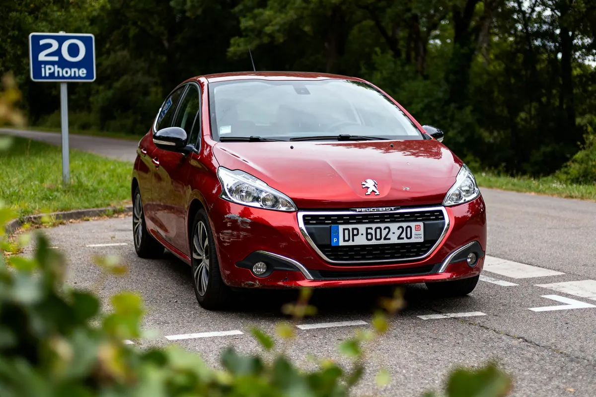 Voiture Peugeot rouge sur route, panneau "20 iPhone", entourée d'arbres, à côté d'un parking.