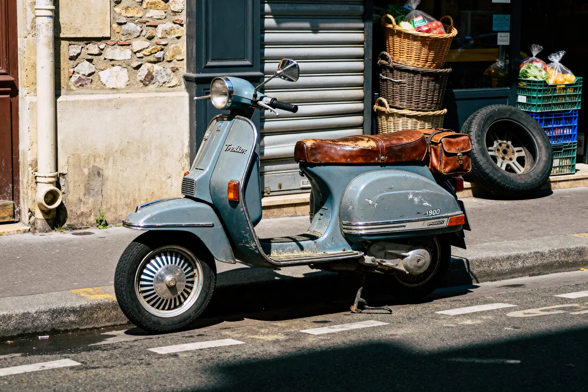 Vieux scooter bleu garé sur trottoir, proche de paniers, devant rideau métallique fermé.