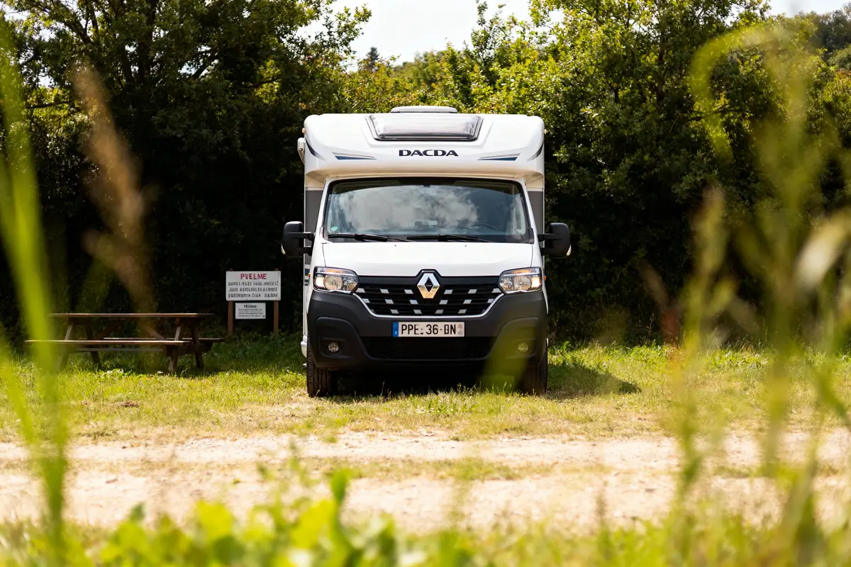 Camping-car blanc Renault sur terrain herbeux, entouré d'arbres, une table de pique-nique à gauche.