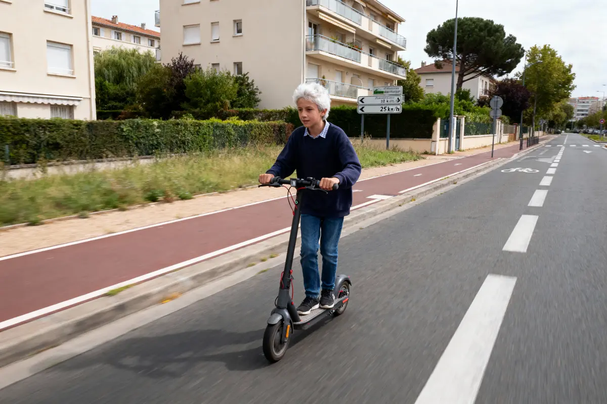 Personne conduisant une trottinette électrique sur une piste cyclable en milieu urbain, bâtiments et verdure en arrière-plan.