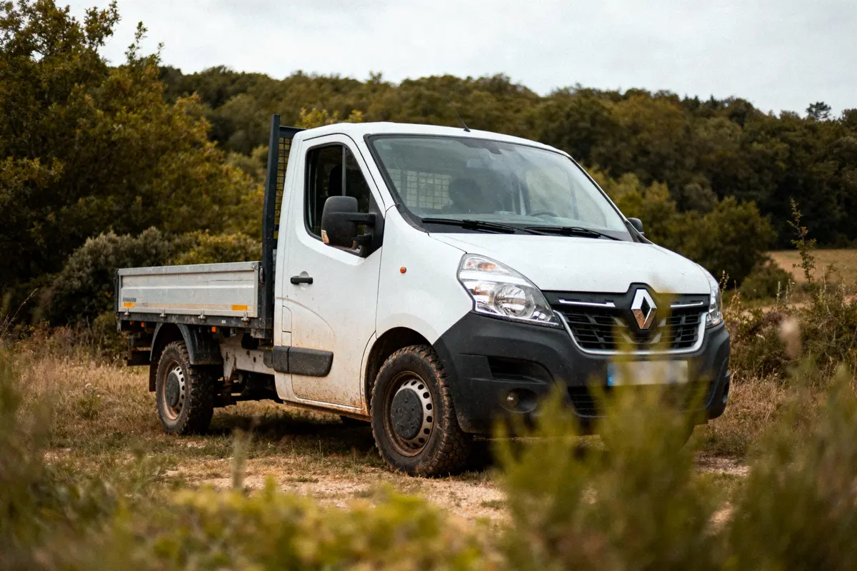 Camionnette blanche Renault garée dans un champ, herbes au premier plan, arbres en arrière-plan.
