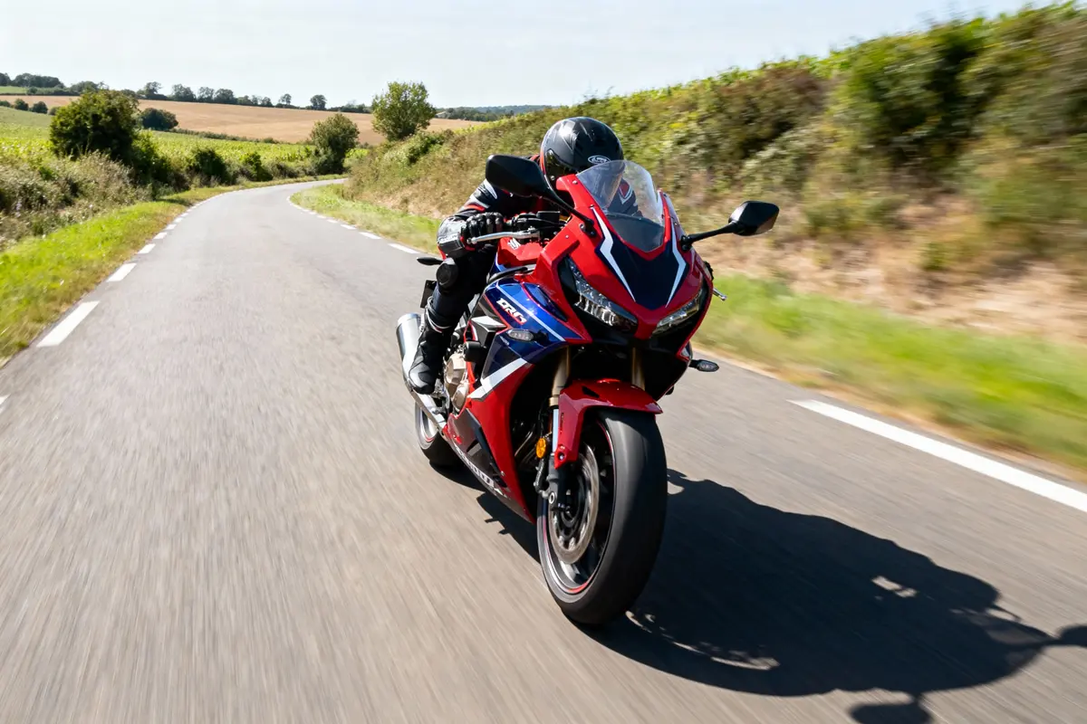 Motard sur une moto rouge, roulant sur une route de campagne sinueuse par temps ensoleillé.