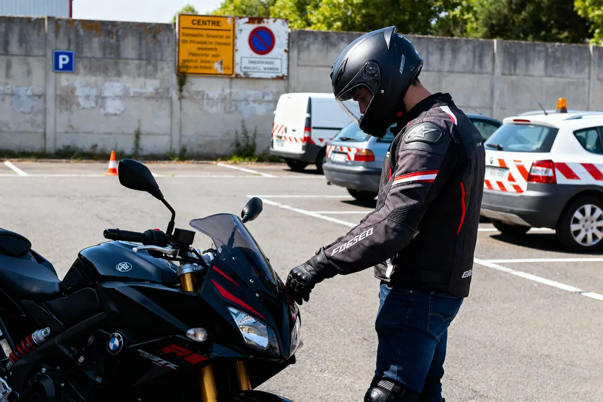 Motard en tenue complète, inspectant une moto sportive noire dans un parking avec panneaux de signalisation en arrière-plan.