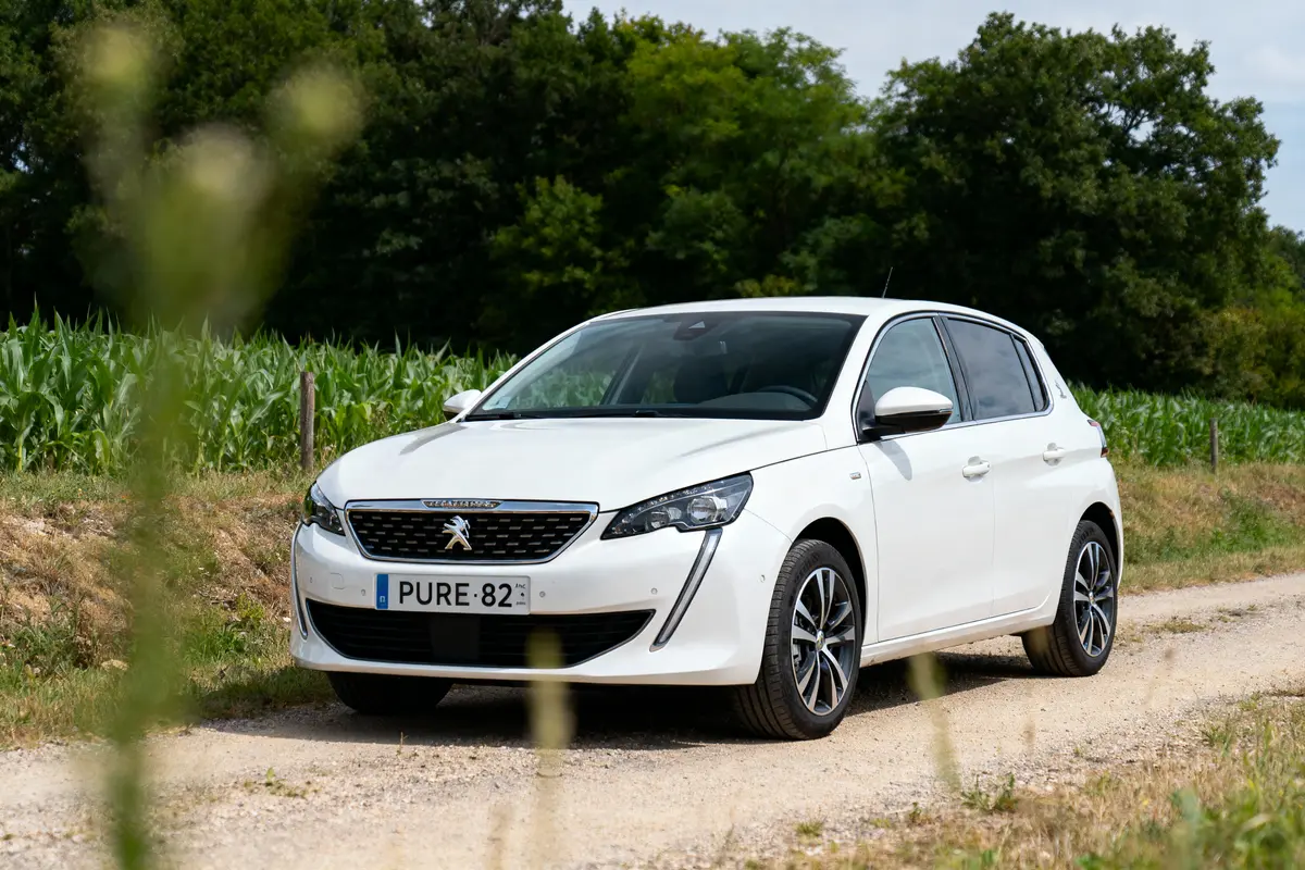 Voiture blanche Peugeot sur chemin de terre, avec végétation et arbres en arrière-plan.