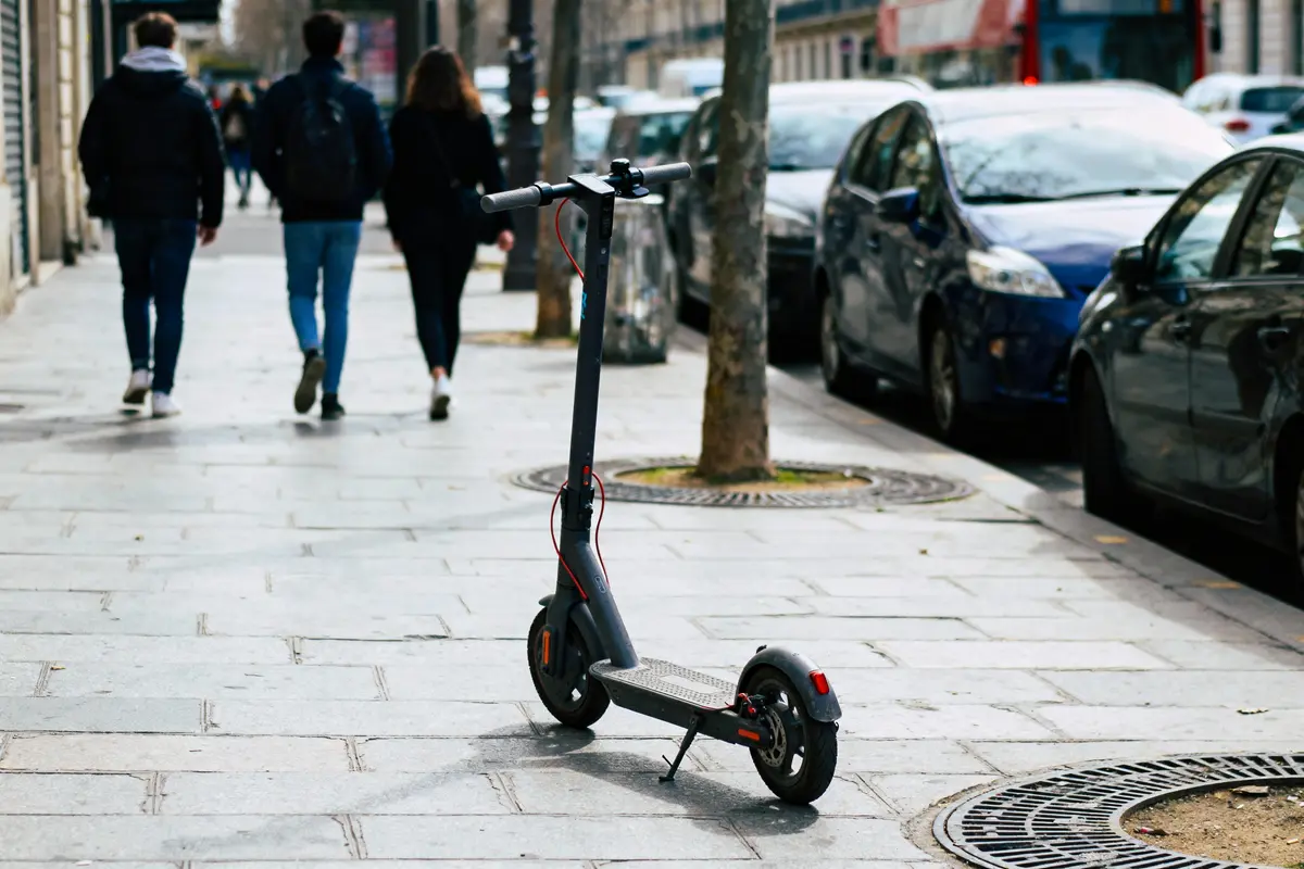 Trottinette électrique sur le trottoir avec voitures garées, arbres et piétons à l'arrière-plan urbain.