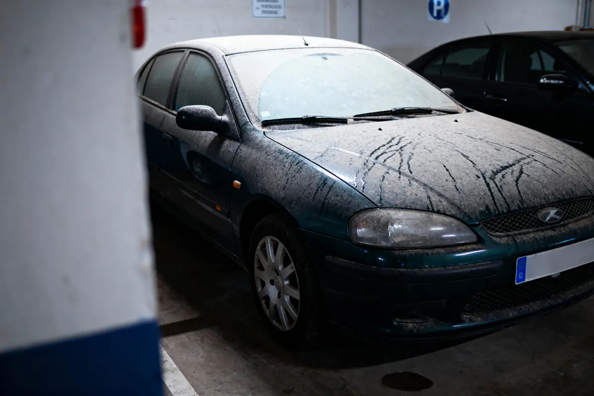 Voiture bleue poussiéreuse garée dans un parking souterrain, partiellement obscurcie par un mur blanc.