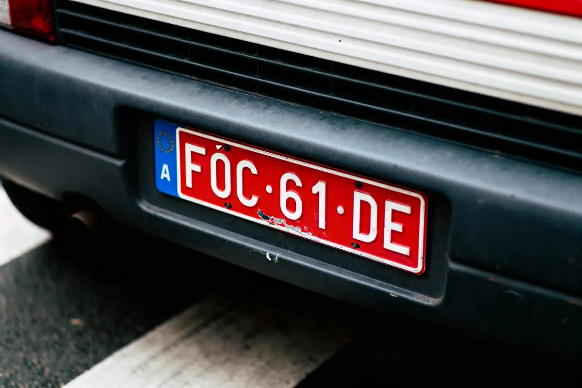 Plaque d'immatriculation rouge sur véhicule, texte “FÓC 61 DE”, bordure blanche et bleue.