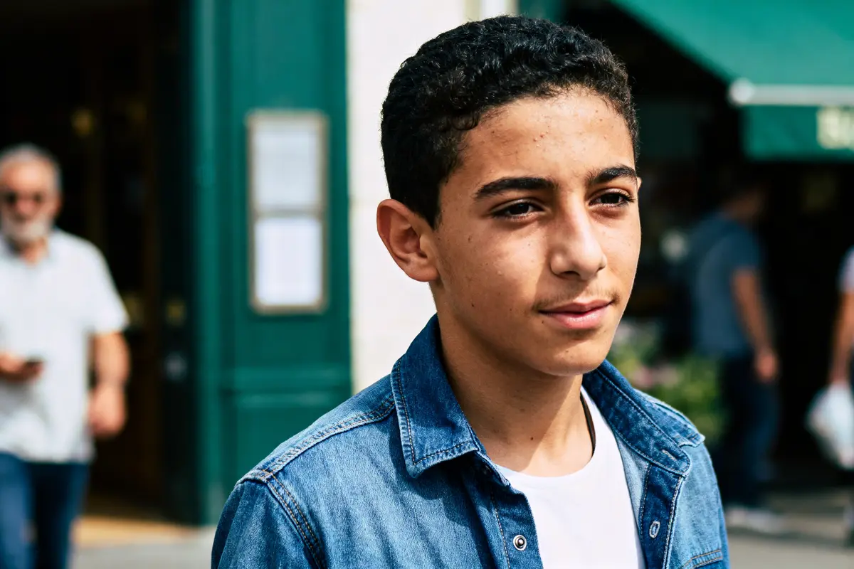Adolescent en chemise en jean, marchant dehors, arrière-plan flou avec d'autres passants.