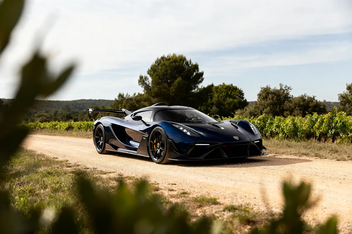 Voiture de sport noire sur chemin de terre, entourée de vignes et arbres, sous ciel légèrement nuageux.