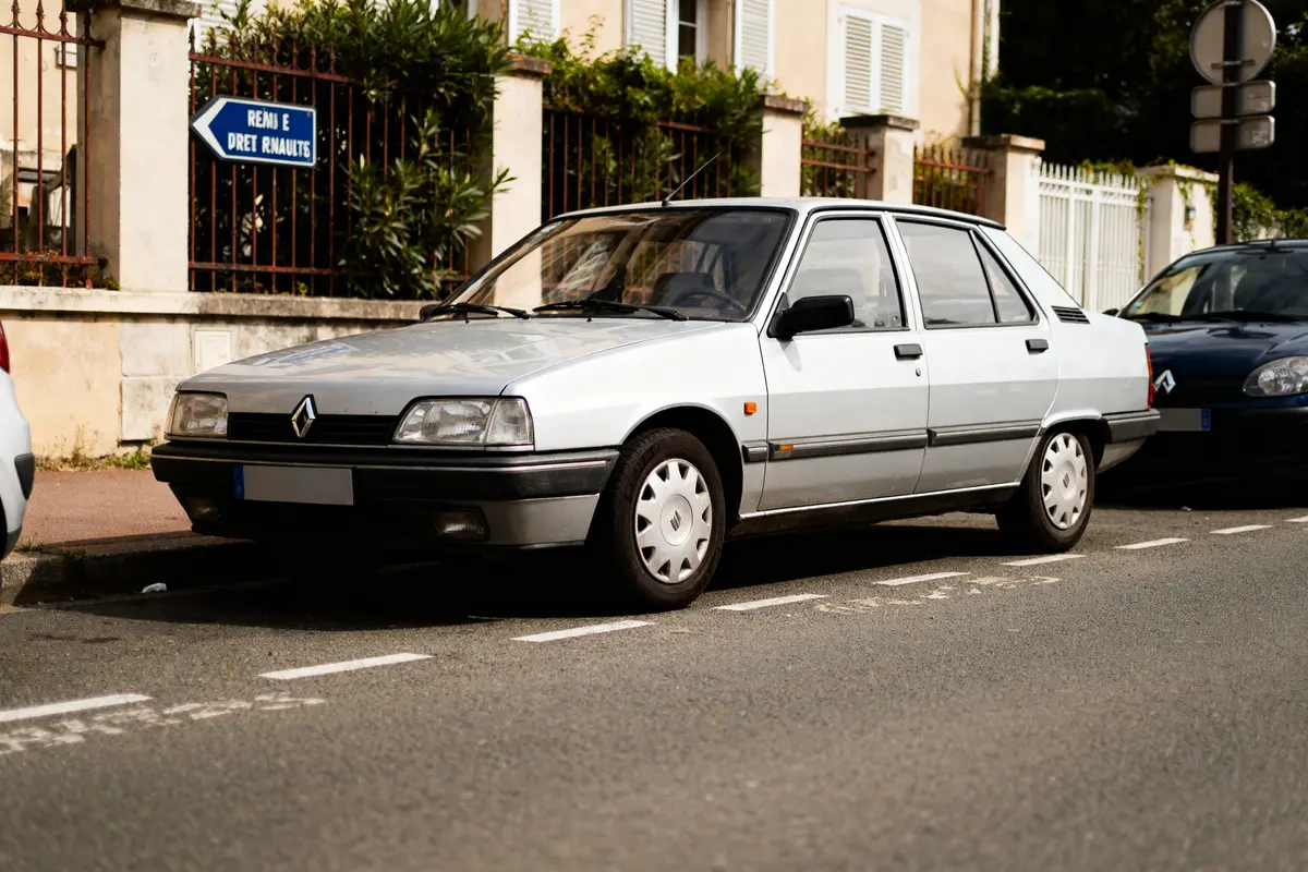 Voiture Renault blanche garée dans une rue, panneau d'indication bleu en arrière-plan, journée ensoleillée.