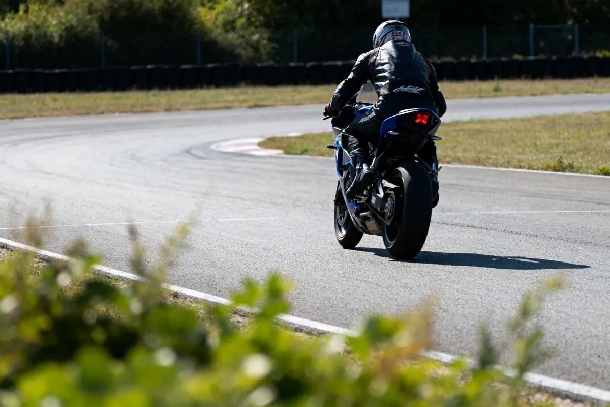 Motard en cuir noir sur circuit, virage sinueux, moto sportive, environnement arboré, herbes au premier plan.