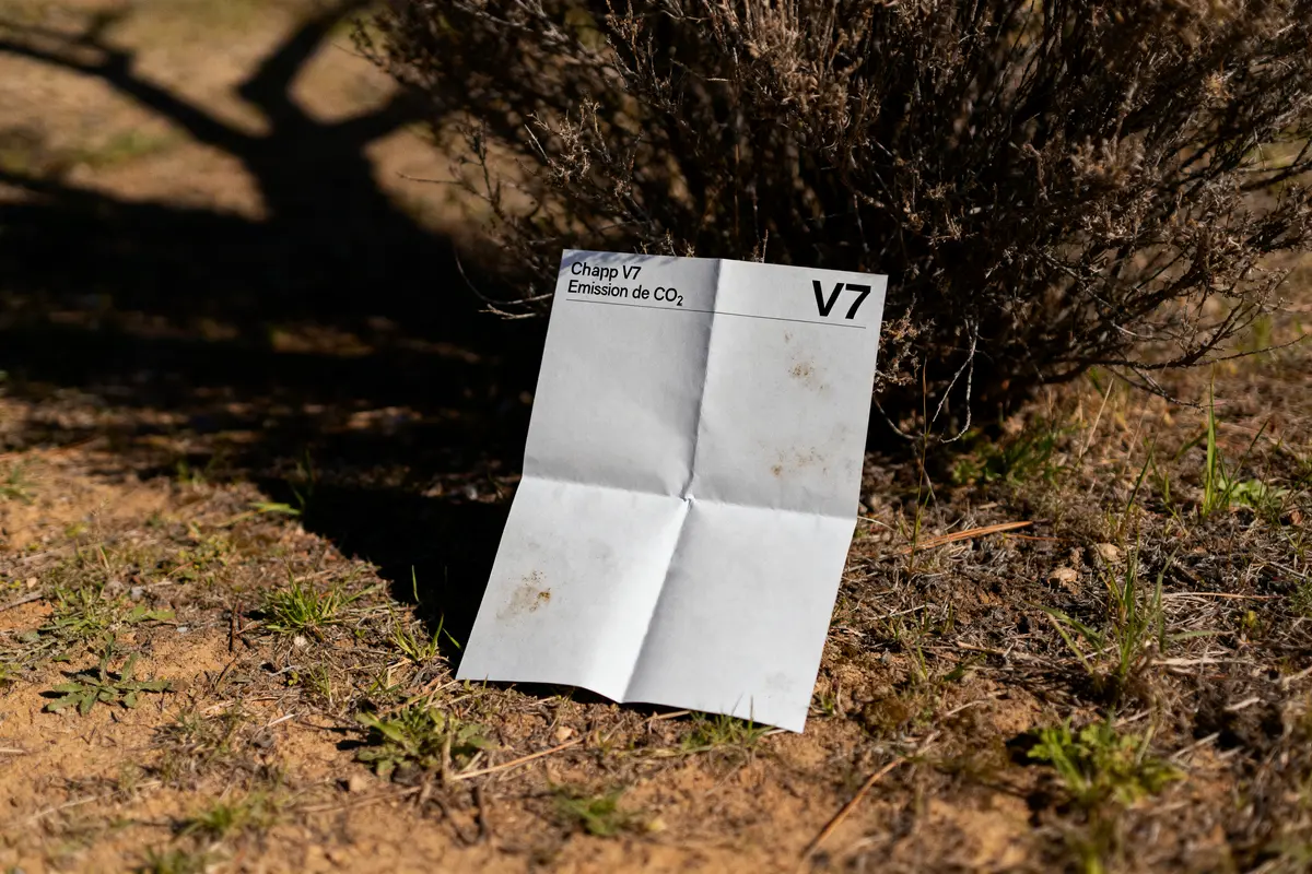 Papier blanc marqué "Chapp V7 Émission de CO₂" posé sur sol terreux près d'un buisson.