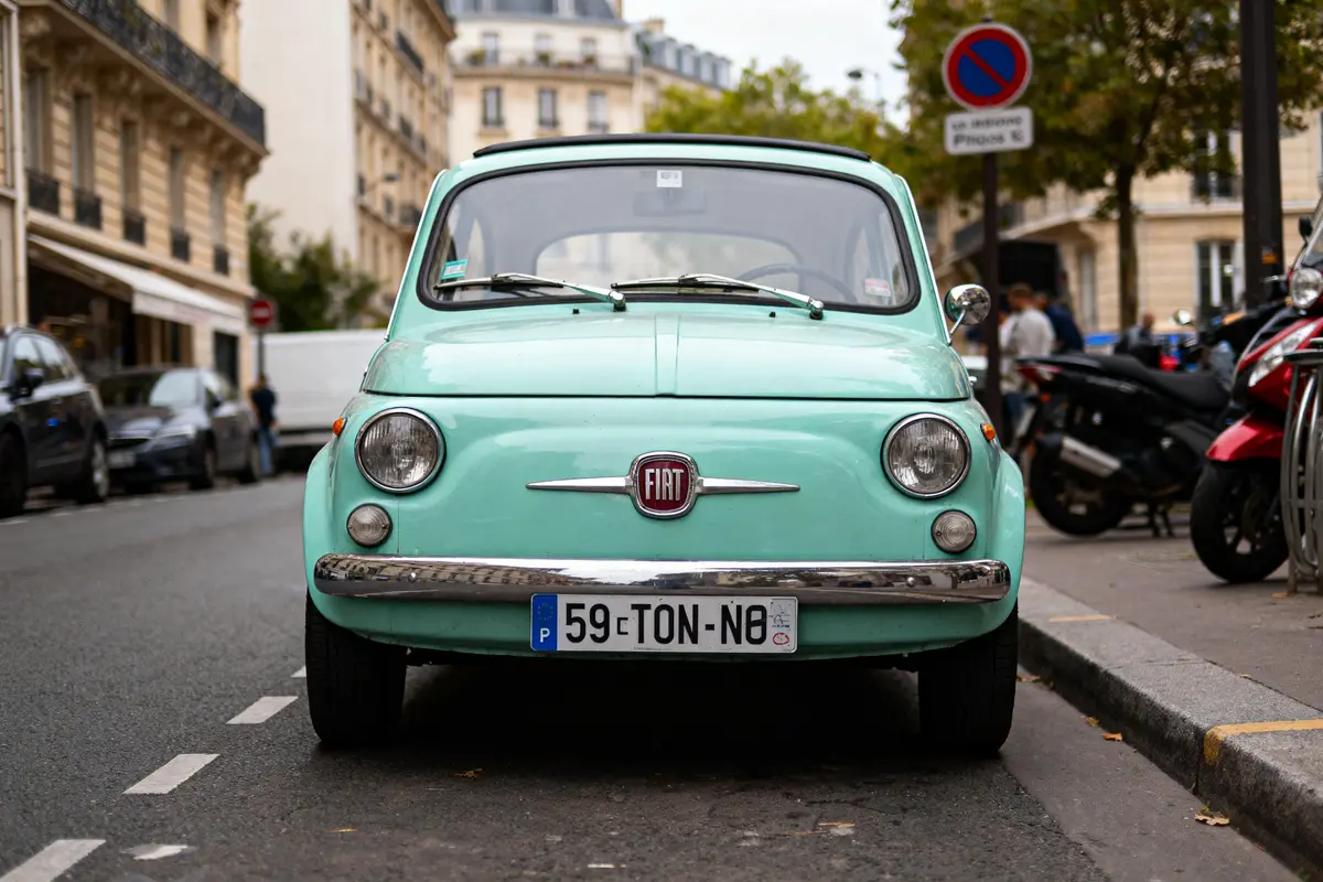 Voiture Fiat 500 vert pastel garée sur rue parisienne, immeubles et motos en arrière-plan.