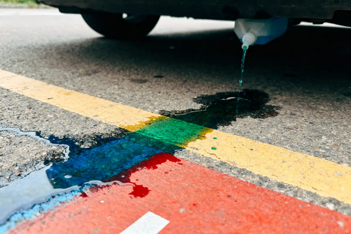 Récipient fuyant sous une voiture, liquide coloré sur chaussée avec bandes jaune et rouge.