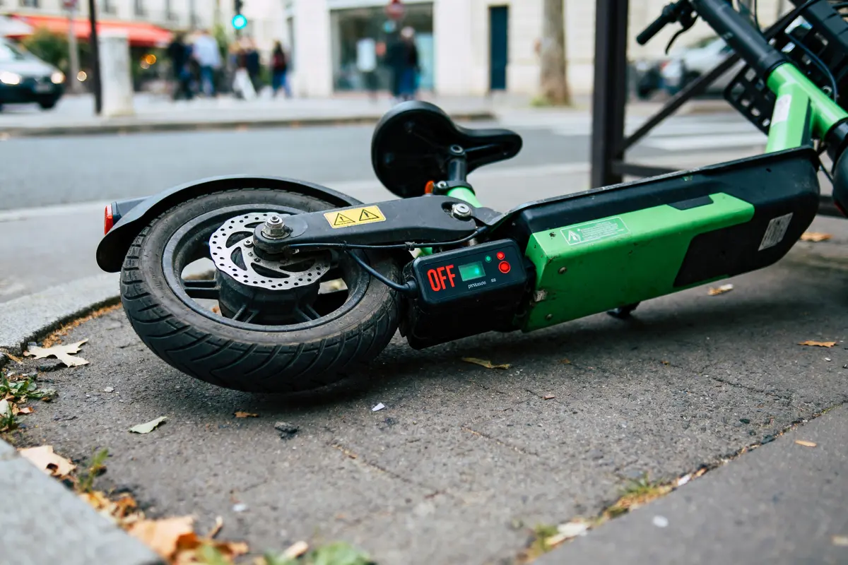 Trottinette verte renversée sur le trottoir, afficheur indiquant "OFF", environnement urbain avec feu tricolore.