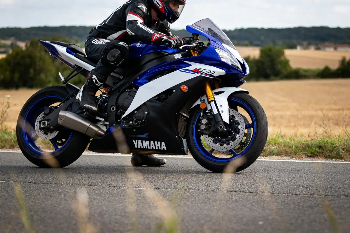 Moto Yamaha R1 bleue sur route, motard en équipement noir, paysage champêtre en arrière-plan.