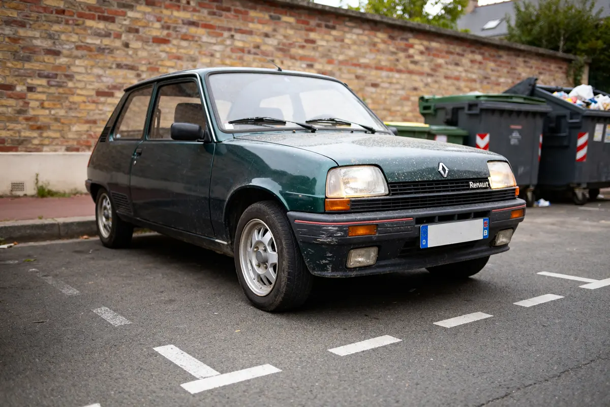 Renault 5 thermique verte garée près de poubelles sur un parking urbain