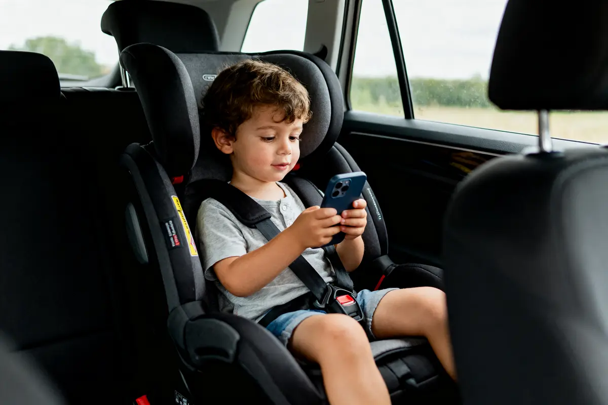 Enfant assis dans un siège auto, utilisant un smartphone, avec ceinture de sécurité, intérieur de voiture visible.
