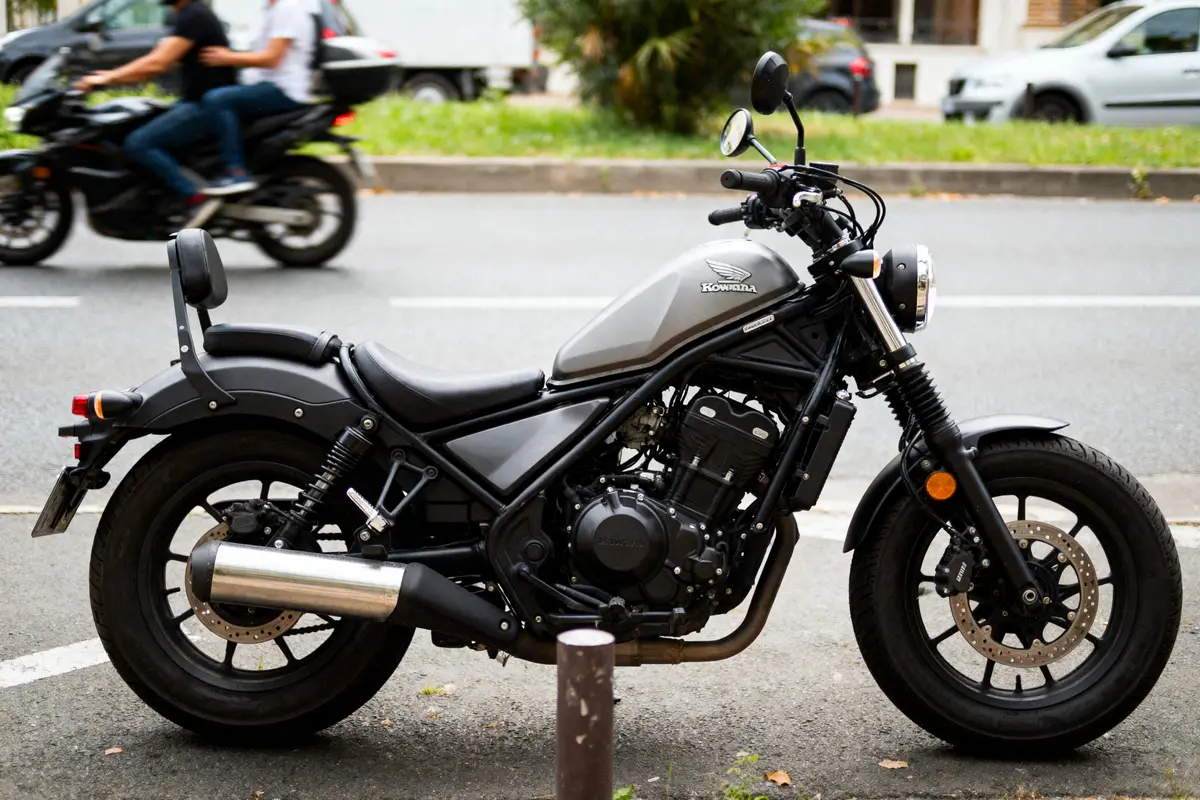 Une moto grise garée sur la route, avec une autre moto en arrière-plan en mouvement.