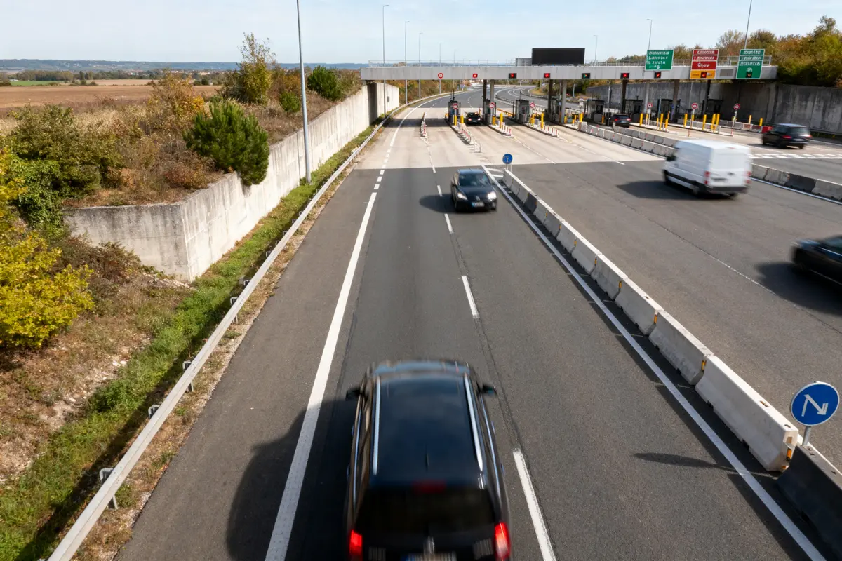 Voitures roulant sur une autoroute près d'un péage, avec barrières et signalisation routière environnante.