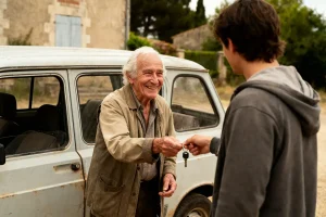 Un homme âgé remet des clés à un jeune devant une vieille voiture.