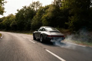 Voiture sportive classique sur route sinueuse, fumée derrière, entourée d'arbres en pleine journée.