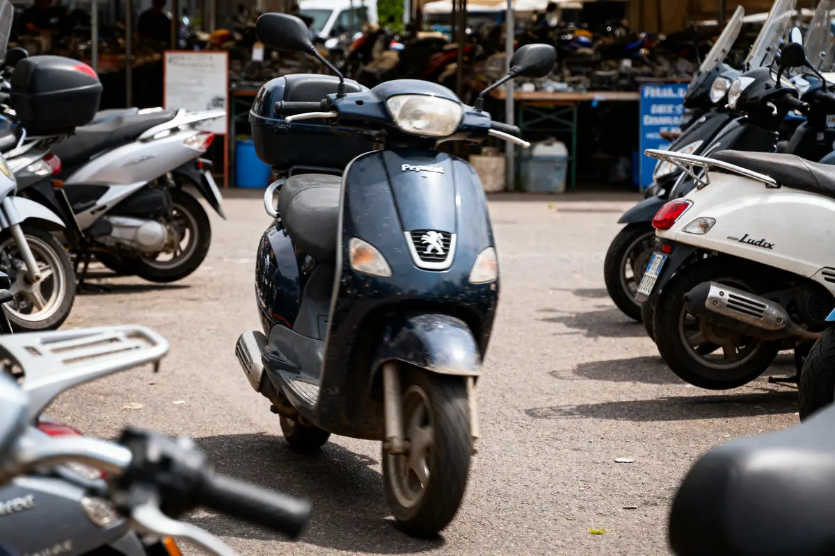 Scooters garés sur un parking, avec un scooter Peugeot en mouvement au centre.