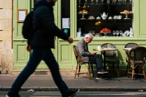 Homme assis à un café parisien, regardant son téléphone, passant piéton flou au premier plan.