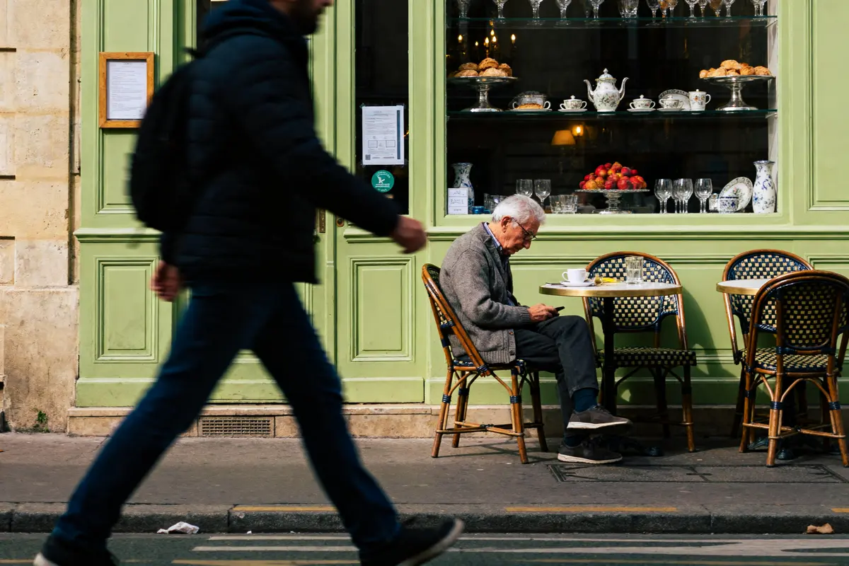 Homme assis à un café parisien, regardant son téléphone, passant piéton flou au premier plan.