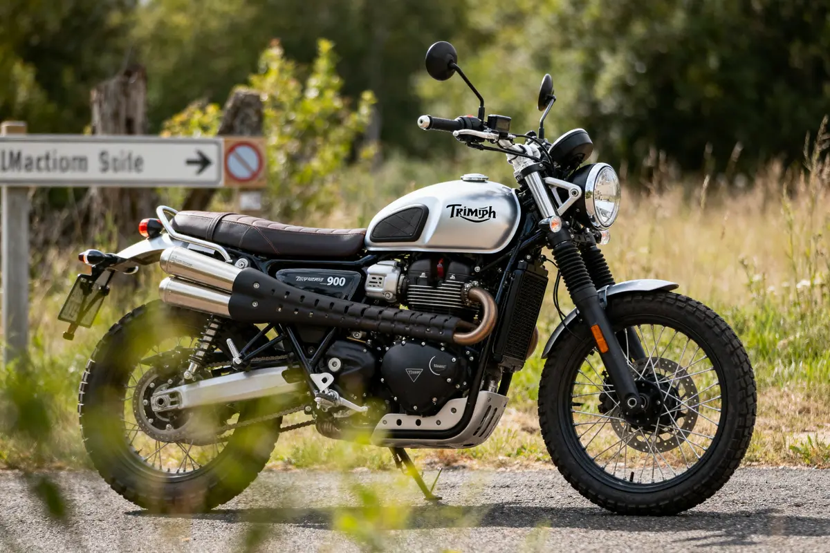 Moto Triumph blanche stationnée sur une route de campagne, végétation en arrière-plan, panneau indicateur.