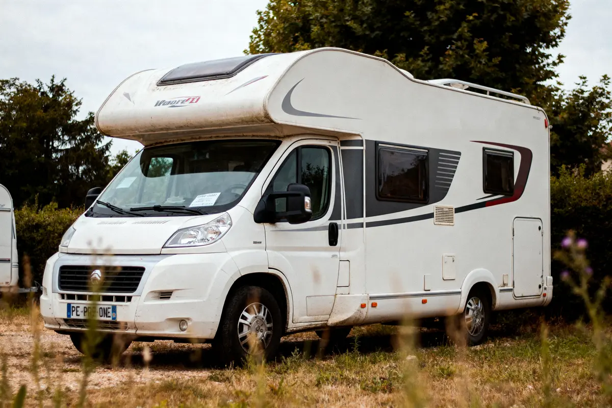 Camping-car blanc stationné sur pelouse, entouré d'arbres, avec motifs gris et noirs sur les côtés.