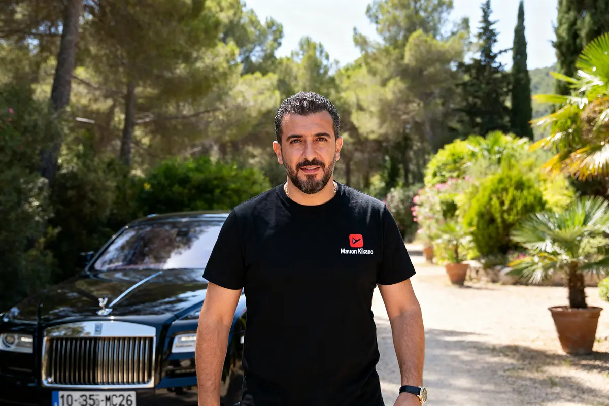Homme en noir devant une voiture de luxe évoquant la fortune de GMK.